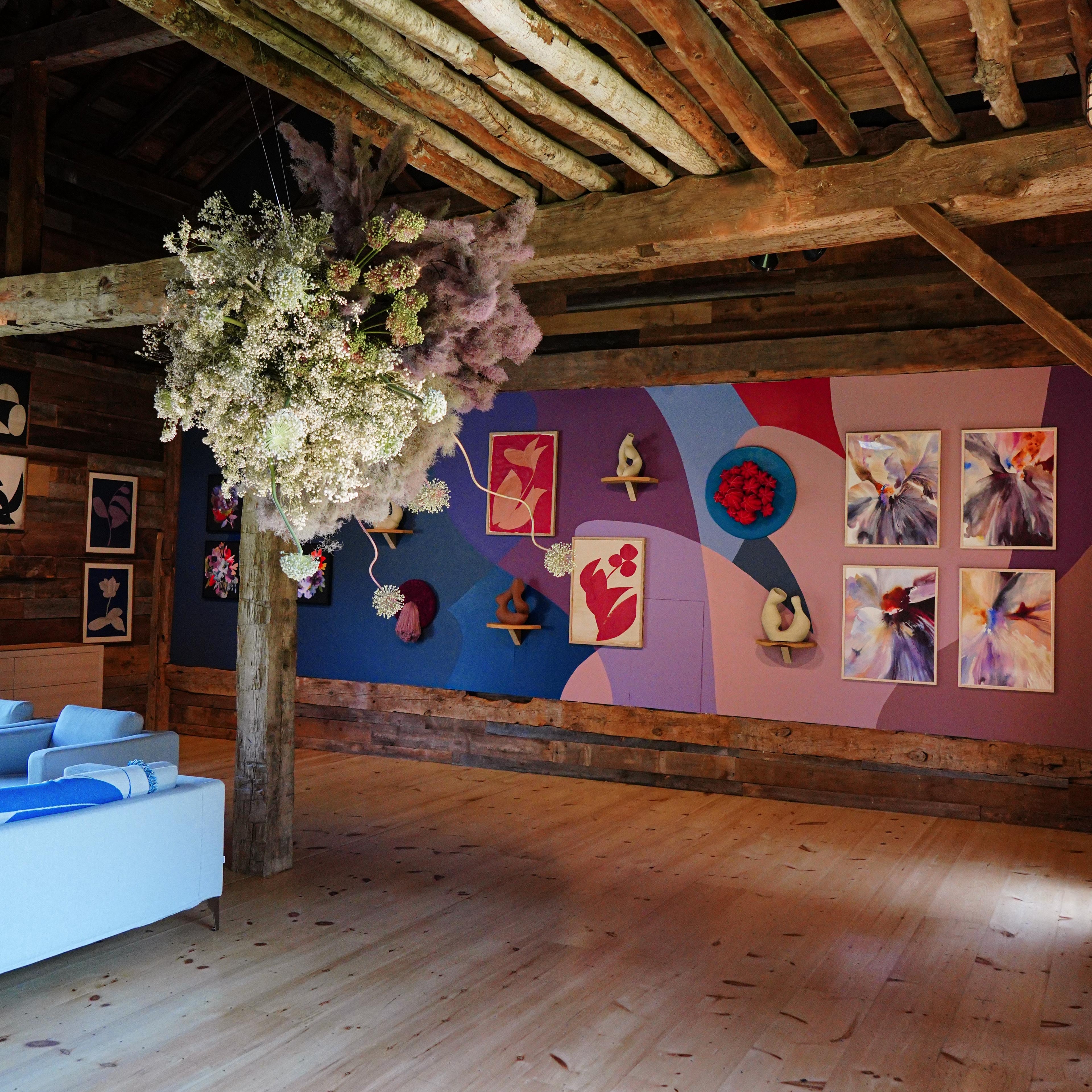 Variety of colorful artwork installed on a bright mural in a rustic showroom with flowers hanging from ceiling 